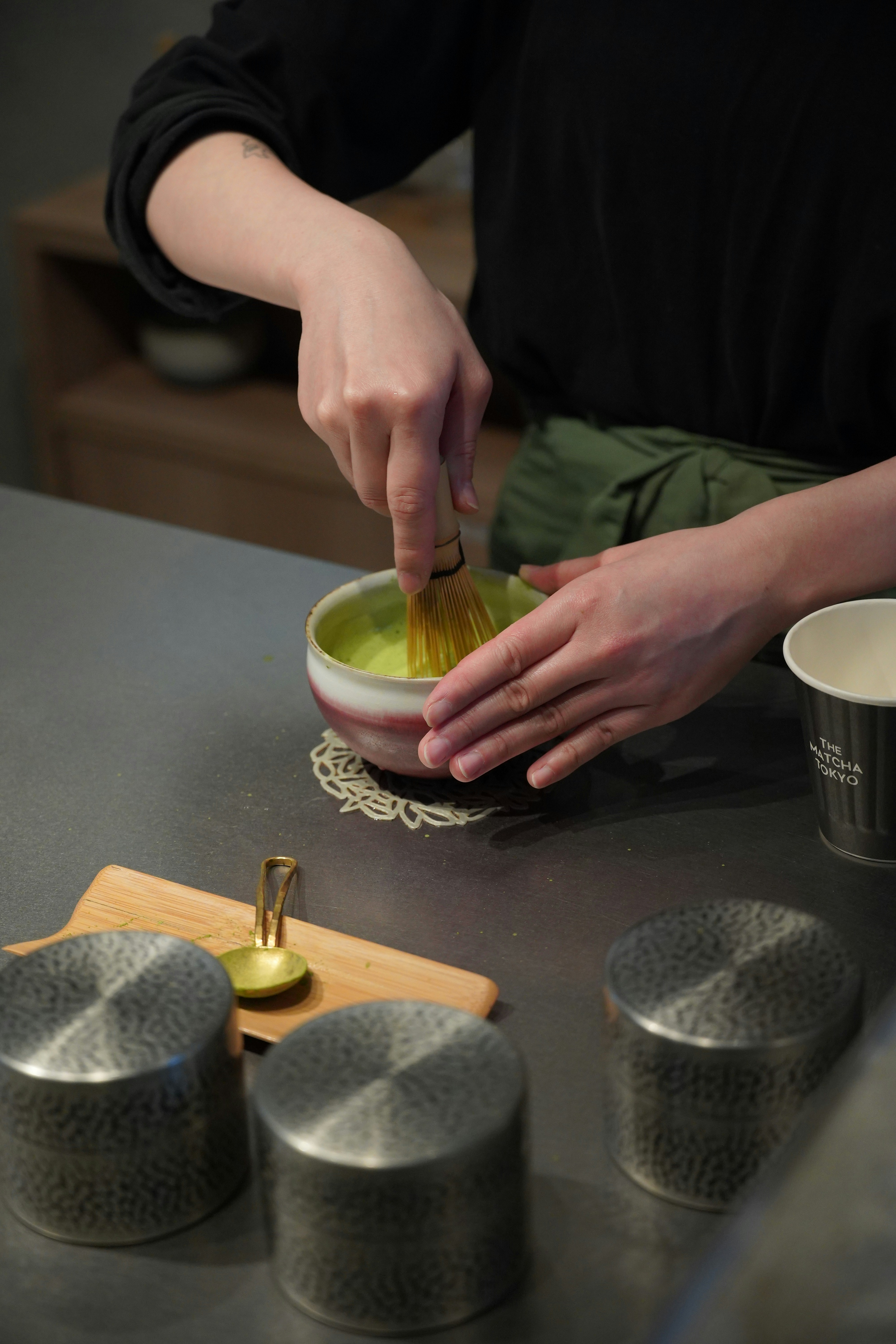 Whisking matcha by hand with a chasen, chashaku, and tea tins nearby